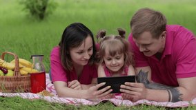 Family weekend picnic. Caucasian daughter with her mother and father lying on grass meadow and play online games on tablet. Child kid watching movies on internet. Chatting, social media network