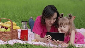 Family weekend at picnic. Caucasian daughter with mother on grass meadow with basket full of fruits play online games on tablet. Child kid watching movies on internet. Chatting, social media network