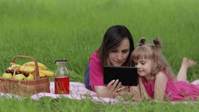 Family weekend at picnic. Caucasian daughter pupil with mother on grass meadow with basket full of fruits learn lessons on tablet. Child kid watching study movies on internet. Distance education