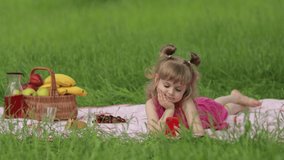 Weekend at picnic. Girl on grass meadow with basket full of fruits play online games on mobile phone. Child kid in pink dress using smartphone social network app for video call, blogging, chatting
