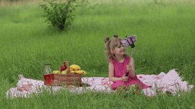 Weekend at picnic. Girl on grass meadow with basket full of fruits makes selfie on mobile phone using selfie stick. Child kid in pink dress using smartphone social network app for video call, blogging