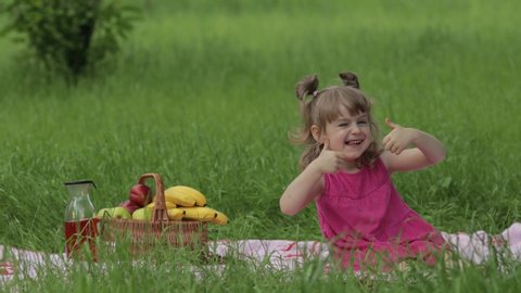 Weekend at picnic. Lovely caucasian girl on green grass meadow with basket full of fruits. Showing thumbs up. Female child kid in pink dress on nature sit on white blanket having fun. Sunny summer