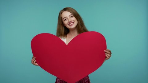 Beautiful happy girl with kind facial expression holding large paper heart and smiling joyfully to camera, imitating heartbeat, demonstrating love care. studio shot isolated on blue background
