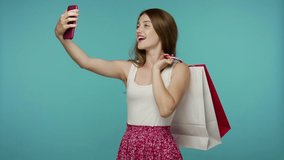 Happy shopper girl in summer dress holding shopping bags and smiling to mobile phone, taking selfie or talking on video call, rejoicing purchase in fashion store. indoor studio shot, blue background