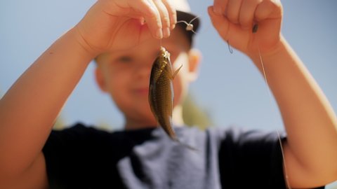Funny little boy holding waving fish on fishing line after catching smiling enjoying hobby close up face of joyful kid having positive emotion at clear blue sky background low angle