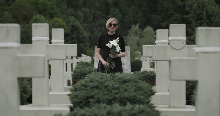 Portrait of young widow looking to camera while standing in row of stone crosses. Woman in dark glasses and clothes holding white lily flower at cemetery. Concept of memorial day.