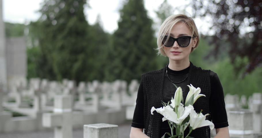 Portrait of sad woman in mourning clothes raising head and looking to camera. Crop view of young widow holding white lily flower while standing at cemetery. Concept of memorial day. Royalty-Free Stock Footage #1055139182