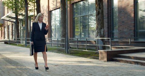 Successful female office worker in formal suit looking at phone screen while walking at business district street. Beautiful businesswoman carrying case and using her phone.