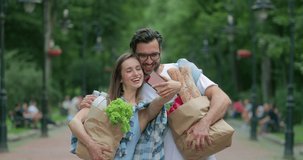 Funny couple doing selfies and posing while walking with paper bags full of food. Cheerful man embracing his wife while they carrying shopping and looking at smartphone screen. Outdoors.