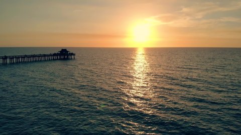 Fly back aerial shot of the silhouette of a fishing pier on the ocean with a colorful and golden sunset. Naples Beach and Fishing Pier at Sunset, Florida. Drone flies back, flying away from the pier.