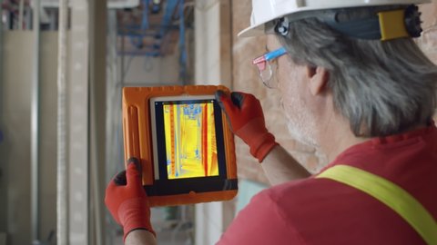 Close up back view of mature foreman in protective hardhat using thermal camera on digital tablet checking thermal insulation strength on construction site