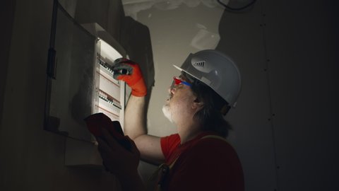 Portrait of mature electrician or foreman in safety uniform and helmet turning on switches in electrician control box holding flashlight on dark construction site