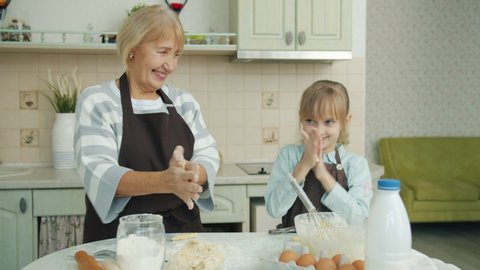 Portrait of old woman and little girl baking in kitchen then making dough mustaches and smiling looking at camera standing near table covered with flour.