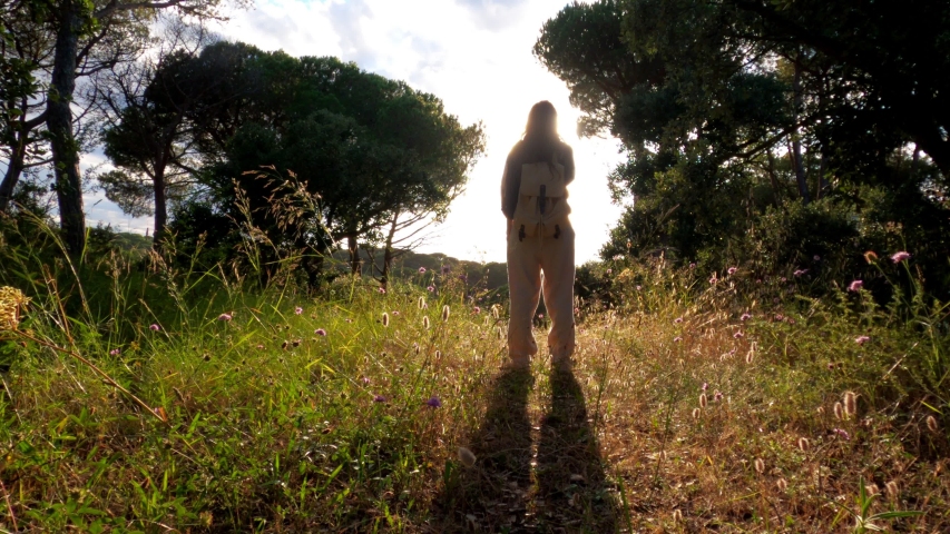 Hiking. A girl with a backpack stands on a forest path and enjoys a beautiful sunset. Golden light in the green forest. Forest hike. Sunny summer day. Walking among the trees and flowers. Holidays.