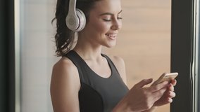 A lovely smiling young woman in a black tracksuit with headphones is listening the music from her smartphone holding and looking on it while standing near a window in a gray studio