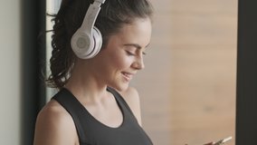 A side-view of a sporty smiling young woman in a black tracksuit is listening to music in headphones standing near the window in a gray studio