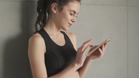 A smiling positive young woman in a black tracksuit is using her smartphone standing near the wall before training in a gray studio in the morning
