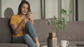 A satisfied pleased young woman is using her smartphone while sitting on the sofa in the living room at home