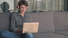 Joyful young pretty woman in headphones talking by videocall on laptop computer while sitting on sofa at home