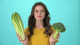 A pretty nice young woman wearing a yellow t-shirt is holding broccoli and chinese cabbage standing isolated over blue background