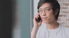 A positive smiling young asian man is talking on his smartphone standing near the window at home