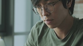 A close-up view of a concentrated young asian freelancer wearing eyeglasses is using his laptop computer while sitting at the table at home