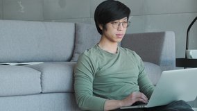 A calm young asian man wearing eyeglasses is using his laptop sitting in the living room at home