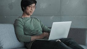 A smiling young asian man wearing eyeglasses is working with his laptop sitting on couch in apartment