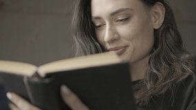Close up view of Happy pretty brunette woman reading book while sitting on chair over grey wall