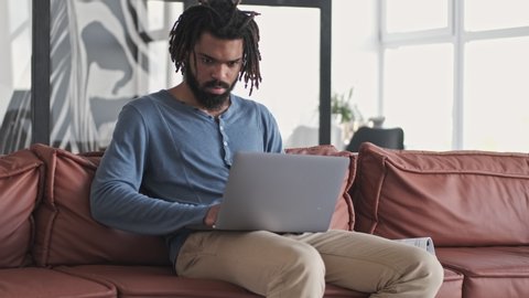 A handsome focused young african american man is using his silver laptop computer in the living room at home