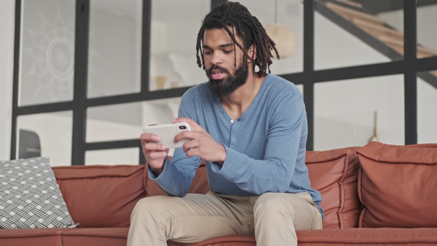 A focused concentrated young african american man is playing games on the smartphone in the living room at home Royalty-Free Stock Footage #1055151488