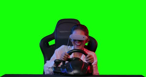 Excited young asian teen gamer wearing vr glasses sitting in the racing seat simulator cockpit with steering wheel and playing in car racing game over green screen background. POV monitor screen view.
