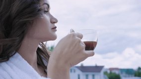 Attractive woman in white coat drinks coffee standing on balcony. Video. Self-sufficient woman is resting in expensive hotel drinking coffee in morning on balcony