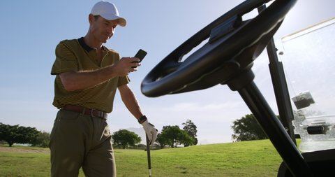 Caucasian male golfer walking on a golf course on a sunny day wearing a cap and golf clothes, using a smartphone and leaning on a golf club, the steering wheel of a golf buggy in the foreground