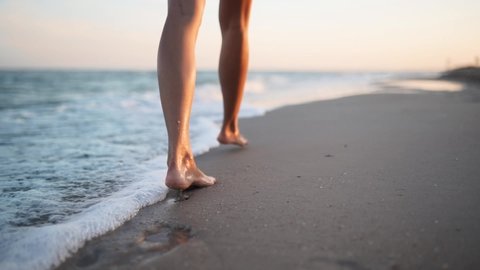 Slim female legs and feet walking along sea water waves on sandy beach. Pretty woman walks at seaside surf. Splashes of water and foam in 120 fps slow motion. Girl after bathing in ocean go on shore.