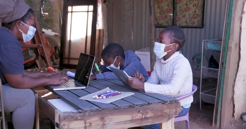 Poverty.Inequality.Black African woman using a laptop and digital tablet to home school her two young children at their dilapidated home during lockdown for Covid-19 Coronavirus pandemic, South Africa