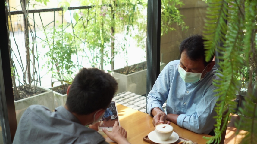 Portrait of a young man teaching his father how to use tablet computer and wearing mask for prevent the spread of the Covid-19. Father and Son using a tablet together at cafe.