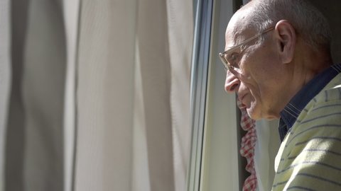 bald old man in glasses looks through window against waving curtains lit by bright sunlight at home slow motion closeup