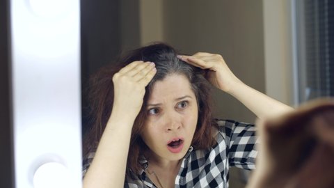 Young Frustrated woman looks in the mirror and plucks a grey hair from her head. Concept photo of young woman getting old