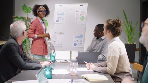 Cheerful African American business lady writing on flipchart, smiling and having discussion with mixed-age diverse team of colleagues while leading corporate training seminar in meeting room