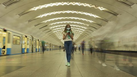 Woman Wearing Mask Standing Still in Underground Metro Station and Using Smartphone during Coronavirus Covid-19 Pandemic. Crowd of People and Passing Trains. Time Lapse