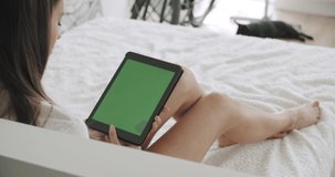 View over head on of caucasian woman lying on bed with black french bulldog at home. Girl holding tablet computer with green screen while watching something. Chroma key. 4k video footage slow motion