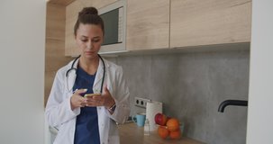 Lunch break in a medical facility. Female doctor in lab coat standing in clinic and scrolling on smartphone screen while kitchen in background. 4k raw video footage slow motion