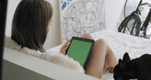 View over head on of caucasian woman lying on bed with black french bulldog at home. Girl holding tablet computer with green screen while watching something. Chroma key. 4k video footage slow motion