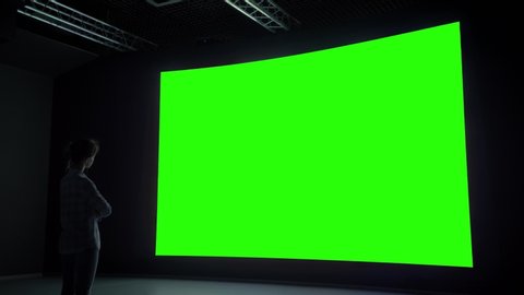 Woman looking at large wall blank interactive green display in dark room of modern technology exhibition or museum. Mock up, green screen, futuristic, template, education, chroma key concept