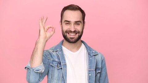 Handsome bearded young man guy 20s years old in denim jacket white t-shirt isolated on pastel pink background studio. People lifestyle concept. Looking camera charming smile showing ok okay gesture