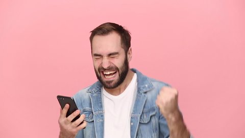 Happy excited young guy 20s years old in denim jacket white t-shirt hold using mobile cell phone just found out great big win news isolated on pastel pink background studio. People lifestyle concept