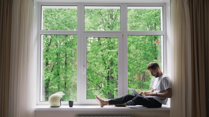 Corona virus - staying and work at home (self-isolation, self-quarantine). A handsome man sits alone in front of a panoramic window with a laptop. Online learning, home work, freelance, 4k