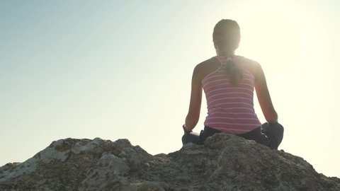 Young relaxed woman sitting outdoors on a big stone enjoying warm summer day. Girl meditating and relaxing on nature at sunset.