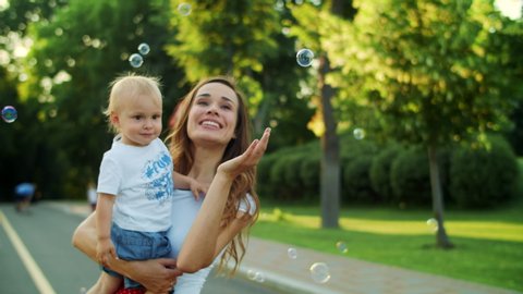 Positive mother holding toddler on hands outdoors. Closeup smiling woman and boy looking at soap bubbles on street. Portrait of happy mother kissing son in summer park. Happy family outdoors